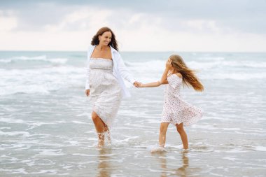 Genç hamile anne ve kızı kumsalda yürüyor. Çocuklu mutlu bir kadın birlikte eğleniyor ve eğleniyor. Çocukluk, hamilelik ve doğa kavramı.