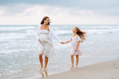 Genç hamile anne ve kızı kumsalda yürüyor. Çocuklu mutlu bir kadın birlikte eğleniyor ve eğleniyor. Çocukluk, hamilelik ve doğa kavramı.