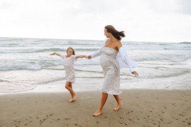 Genç hamile anne ve kızı kumsalda yürüyor. Çocuklu mutlu bir kadın birlikte eğleniyor ve eğleniyor. Çocukluk, hamilelik ve doğa kavramı.