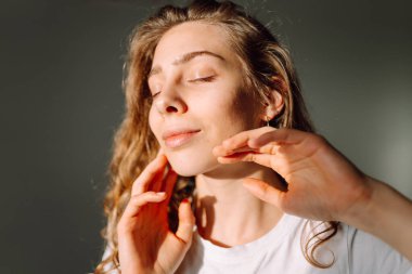 Sıcak bir odada otururken yüzüne dokunan sevimli bir kadının portresi. Güneş ışınlarının ve doğal güzelliğin tadını çıkaran güzel bir kadın. Doğal güzellik, dermatoloji ve kozmetoloji kavramı.