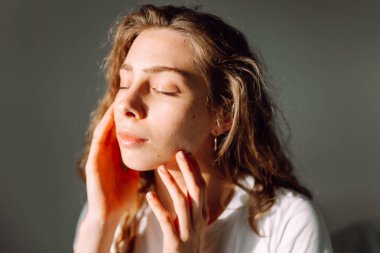 Sıcak bir odada otururken yüzüne dokunan sevimli bir kadının portresi. Güneş ışınlarının ve doğal güzelliğin tadını çıkaran güzel bir kadın. Doğal güzellik, dermatoloji ve kozmetoloji kavramı.