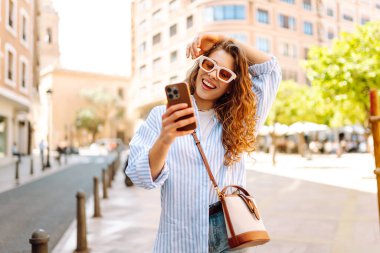 Güzel bir turist, güneşli bir caddede kaldırımda elinde telefonla oturur. Telefonu olan genç bir turist blog yazıyor ve şehir yürüyüşünden zevk alıyor. Blogculuk, tatil ve turizm kavramı.