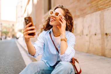 Güzel bir turist, güneşli bir caddede kaldırımda elinde telefonla oturur. Telefonu olan genç bir turist blog yazıyor ve şehir yürüyüşünden zevk alıyor. Blogculuk, tatil ve turizm kavramı.