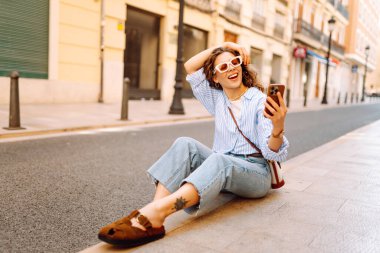 Güzel bir turist, güneşli bir caddede kaldırımda elinde telefonla oturur. Telefonu olan genç bir turist blog yazıyor ve şehir yürüyüşünden zevk alıyor. Blogculuk, tatil ve turizm kavramı.