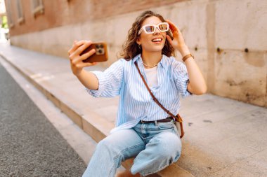 Güzel bir turist, güneşli bir caddede kaldırımda elinde telefonla oturur. Telefonu olan genç bir turist blog yazıyor ve şehir yürüyüşünden zevk alıyor. Blogculuk, tatil ve turizm kavramı.