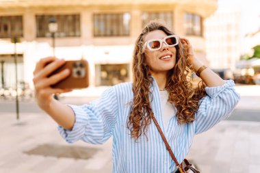 Güzel bir turist, güneşli bir caddede kaldırımda elinde telefonla oturur. Telefonu olan genç bir turist blog yazıyor ve şehir yürüyüşünden zevk alıyor. Blogculuk, tatil ve turizm kavramı.