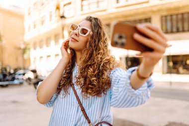 Güzel bir turist, güneşli bir caddede kaldırımda elinde telefonla oturur. Telefonu olan genç bir turist blog yazıyor ve şehir yürüyüşünden zevk alıyor. Blogculuk, tatil ve turizm kavramı.