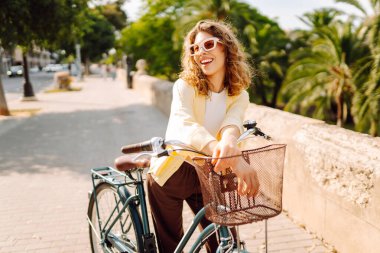 Güneşli şehir sokaklarında bisiklet süren mutlu bir kadın. Parlak elbiseli güzel bir kadın dışarıda bisiklet sürerek eğleniyor. Şehir, eğlenceli konsept. Etkin yaşam biçimi.