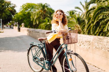 Güneşli şehir sokaklarında bisiklet süren mutlu bir kadın. Parlak elbiseli güzel bir kadın dışarıda bisiklet sürerek eğleniyor. Şehir, eğlenceli konsept. Etkin yaşam biçimi.