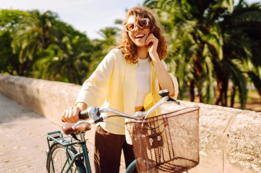 Güneşli şehir sokaklarında bisiklet süren mutlu bir kadın. Parlak elbiseli güzel bir kadın dışarıda bisiklet sürerek eğleniyor. Şehir, eğlenceli konsept. Etkin yaşam biçimi.