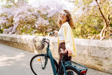 Güneşli şehir sokaklarında bisiklet süren mutlu bir kadın. Parlak elbiseli güzel bir kadın dışarıda bisiklet sürerek eğleniyor. Şehir, eğlenceli konsept. Etkin yaşam biçimi.