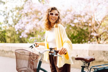 Güneşli şehir sokaklarında bisiklet süren mutlu bir kadın. Parlak elbiseli güzel bir kadın dışarıda bisiklet sürerek eğleniyor. Şehir, eğlenceli konsept. Etkin yaşam biçimi.