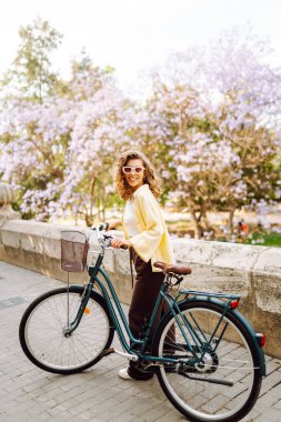 Güneşli şehir sokaklarında bisiklet süren mutlu bir kadın. Parlak elbiseli güzel bir kadın dışarıda bisiklet sürerek eğleniyor. Şehir, eğlenceli konsept. Etkin yaşam biçimi.