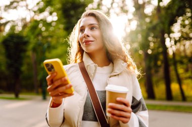 Günlük kıyafetlerle gülümseyen bir kadın güneşli bir parkta elinde telefon ve bir fincan kahveyle yürüyor. Genç bir kadın dışarıda sohbet ediyor ya da blog yazıyor. Güzellik, teknoloji ve yürüyüş konsepti.