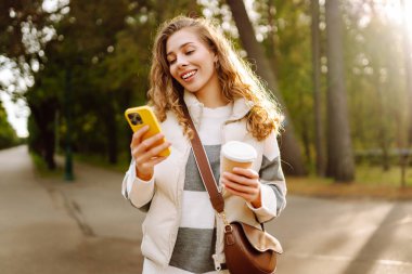 Günlük kıyafetlerle gülümseyen bir kadın güneşli bir parkta elinde telefon ve bir fincan kahveyle yürüyor. Genç bir kadın dışarıda sohbet ediyor ya da blog yazıyor. Güzellik, teknoloji ve yürüyüş konsepti.