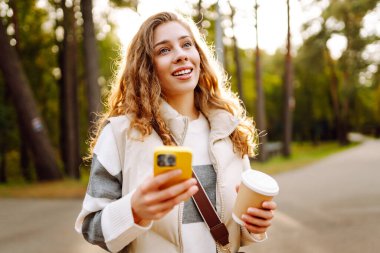 Günlük kıyafetlerle gülümseyen bir kadın güneşli bir parkta elinde telefon ve bir fincan kahveyle yürüyor. Genç bir kadın dışarıda sohbet ediyor ya da blog yazıyor. Güzellik, teknoloji ve yürüyüş konsepti.