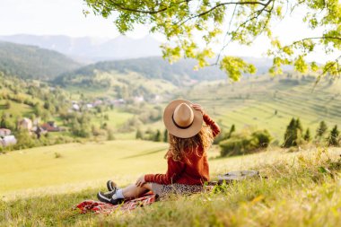 Şapkalı yürüyüşçü kadın yeşil tepedeki battaniyeye oturmuş güneşli manzaranın tadını çıkarıyor. Güzel bir kadın özgürlüğü hisseder ve tepelerde yürür. Seyahat, eğlence ve özgürlük kavramı.