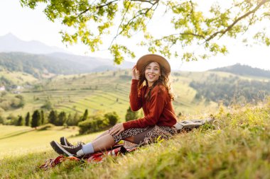 Şapkalı yürüyüşçü kadın yeşil tepedeki battaniyeye oturmuş güneşli manzaranın tadını çıkarıyor. Güzel bir kadın özgürlüğü hisseder ve tepelerde yürür. Seyahat, eğlence ve özgürlük kavramı.