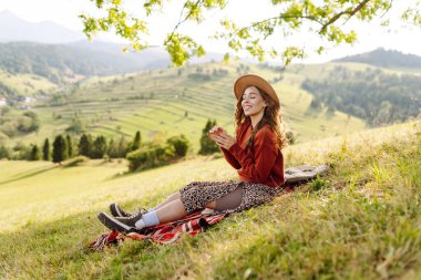 Şapkalı yürüyüşçü kadın yeşil tepedeki battaniyeye oturmuş güneşli manzaranın tadını çıkarıyor. Güzel bir kadın özgürlüğü hisseder ve tepelerde yürür. Seyahat, eğlence ve özgürlük kavramı.