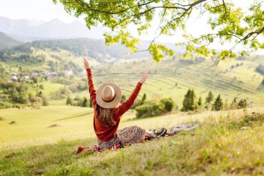 Şapkalı yürüyüşçü kadın yeşil tepedeki battaniyeye oturmuş güneşli manzaranın tadını çıkarıyor. Güzel bir kadın özgürlüğü hisseder ve tepelerde yürür. Seyahat, eğlence ve özgürlük kavramı.