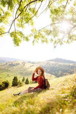 Şapkalı yürüyüşçü kadın yeşil tepedeki battaniyeye oturmuş güneşli manzaranın tadını çıkarıyor. Güzel bir kadın özgürlüğü hisseder ve tepelerde yürür. Seyahat, eğlence ve özgürlük kavramı.