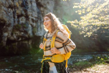 Dağ nehri yakınlarında sırt çantasıyla gezen genç bayan gezgin dağ manzarasının tadını çıkarıyor. Kıvırcık kadın doğada kendini rahat hisseder ve dağlarda ormanda yürür. Seyahat konsepti, etkinlik.