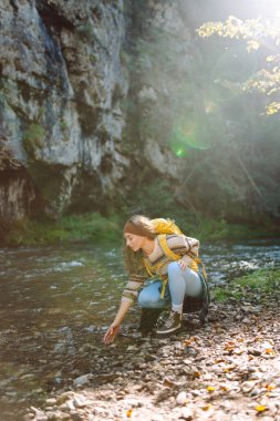 Dağ nehri yakınlarında sırt çantasıyla gezen genç bayan gezgin dağ manzarasının tadını çıkarıyor. Kıvırcık kadın doğada kendini rahat hisseder ve dağlarda ormanda yürür. Seyahat konsepti, etkinlik.