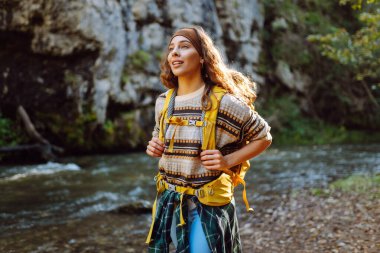 Dağ nehri yakınlarında sırt çantasıyla gezen genç bayan gezgin dağ manzarasının tadını çıkarıyor. Kıvırcık kadın doğada kendini rahat hisseder ve dağlarda ormanda yürür. Seyahat konsepti, etkinlik.