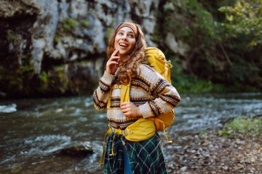 Dağ nehri yakınlarında sırt çantasıyla gezen genç bayan gezgin dağ manzarasının tadını çıkarıyor. Kıvırcık kadın doğada kendini rahat hisseder ve dağlarda ormanda yürür. Seyahat konsepti, etkinlik.