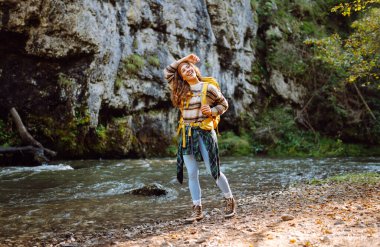 Dağ nehri yakınlarında sırt çantasıyla gezen genç bayan gezgin dağ manzarasının tadını çıkarıyor. Kıvırcık kadın doğada kendini rahat hisseder ve dağlarda ormanda yürür. Seyahat konsepti, etkinlik.
