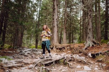 Dağlarda yürüyen kayalık bir patikada renkli bir sırt çantasıyla kadın yürüyüşçü. Yoğun bir ormanda dağ manzarasından hoşlanan sevimli bir kadın. Macera, yürüyüş ve doğa konsepti.