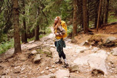 Dağlarda yürüyen kayalık bir patikada renkli bir sırt çantasıyla kadın yürüyüşçü. Yoğun bir ormanda dağ manzarasından hoşlanan sevimli bir kadın. Macera, yürüyüş ve doğa konsepti.