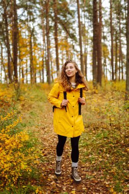 Genç bir kadın sonbahar parkında eğleniyor, sarı yapraklar saçıyor. Parlak yağmurluklu kadın gezgin gün batımında ormanda güzel bir yolda yürüyor. Mevsimlik kavramı, sonbahar