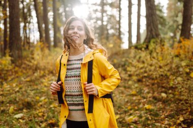 Genç bir kadın sonbahar parkında eğleniyor, sarı yapraklar saçıyor. Parlak yağmurluklu kadın gezgin gün batımında ormanda güzel bir yolda yürüyor. Mevsimlik kavramı, sonbahar