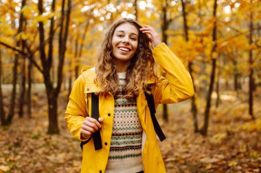 Genç bir kadın sonbahar parkında eğleniyor, sarı yapraklar saçıyor. Parlak yağmurluklu kadın gezgin gün batımında ormanda güzel bir yolda yürüyor. Mevsimlik kavramı, sonbahar