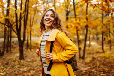 Genç bir kadın sonbahar parkında eğleniyor, sarı yapraklar saçıyor. Parlak yağmurluklu kadın gezgin gün batımında ormanda güzel bir yolda yürüyor. Mevsimlik kavramı, sonbahar