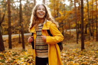 Genç bir kadın sonbahar parkında eğleniyor, sarı yapraklar saçıyor. Parlak yağmurluklu kadın gezgin gün batımında ormanda güzel bir yolda yürüyor. Mevsimlik kavramı, sonbahar