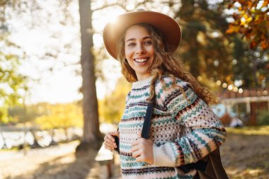 Şapkalı genç bir kadın gülerek sonbahar parkında güneşli bir günün tadını çıkarıyor. Dışarıda yürüyen mutlu bir kadın. Aktif yaşam tarzı. Yürüyüş ve doğa konsepti.