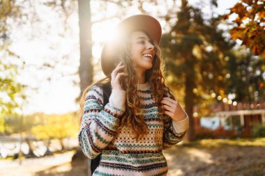 Şapkalı genç bir kadın gülerek sonbahar parkında güneşli bir günün tadını çıkarıyor. Dışarıda yürüyen mutlu bir kadın. Aktif yaşam tarzı. Yürüyüş ve doğa konsepti.