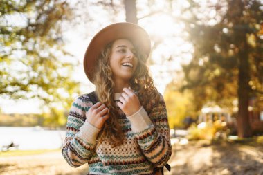 Şapkalı genç bir kadın gülerek sonbahar parkında güneşli bir günün tadını çıkarıyor. Dışarıda yürüyen mutlu bir kadın. Aktif yaşam tarzı. Yürüyüş ve doğa konsepti.