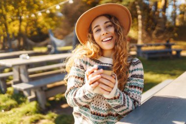 Ahşap bir masada şapkalı neşeli bir kadın ve bir bardak sıcak içecekle sonbahar parkının tadını çıkarıyor. Genç bir kadın güneşli bir günde eğleniyor ve yürüyor. Doğa kavramı, içkiler ve rahatlama.