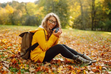 Parlak yağmurluklu kadın gezgin, sonbahar yapraklarıyla çevrili termostan sıcak çay içerek yeşil bir çayırda oturur. Şirin bir kadın güneşli bir sonbahar gününün tadını çıkarıyor. Hafta sonu ve macera konsepti.