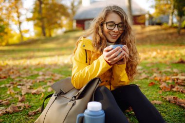 Parlak yağmurluklu kadın gezgin, sonbahar yapraklarıyla çevrili termostan sıcak çay içerek yeşil bir çayırda oturur. Şirin bir kadın güneşli bir sonbahar gününün tadını çıkarıyor. Hafta sonu ve macera konsepti.