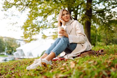Güzel bir kadın gölün kenarında bir battaniyeye oturup sıcak bir içecek içer. Parkta poz veren genç bir kadın. Rahatlama, doğa konsepti..