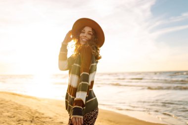 Şapkalı neşeli kadın güneşli sahilde eğleniyor ve yürüyor. Özgürlüğü hisseden ve deniz manzarasının tadını çıkaran güzel bir kadın. Özgürlük, rahatlama konsepti. Yaşam biçimi.