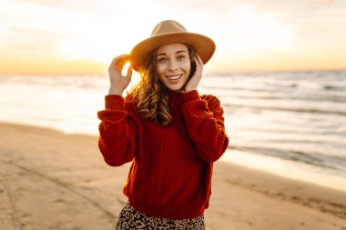 Şapkalı neşeli kadın güneşli sahilde eğleniyor ve yürüyor. Özgürlüğü hisseden ve deniz manzarasının tadını çıkaran güzel bir kadın. Özgürlük, rahatlama konsepti. Yaşam biçimi.