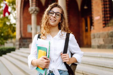 Üniversite avlusunda güzel bir bayan öğrenci elinde defterler ve kitaplarla kampüsün arka planına karşı. Üniversitede okuduktan sonra gözlüklü genç bir kadın. Eğitim kavramı.