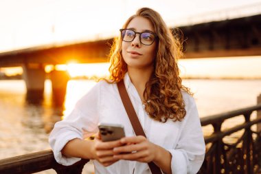 Kıvırcık saçlı, gözlüklü, telefonlu genç bir kadın şehrin seddindeki günbatımının tadını çıkarıyor. Telefonuyla mesaj atan, dışarıda çalışan hoş bir kadın. Teknoloji ve eğlence kavramı.