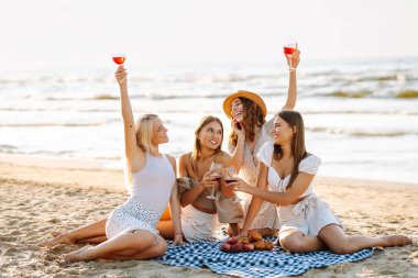 Deniz kıyısında piknik yapan neşeli kadınlar birlikte vakit geçiriyorlar, doğada dinleniyorlar. Dört kız arkadaş eğleniyor, şarap içiyor ve açık havada gün batımıyla buluşuyorlar. Dostluk kavramı, kutlama.