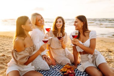 Deniz kıyısında piknik yapan neşeli kadınlar birlikte vakit geçiriyorlar, doğada dinleniyorlar. Dört kız arkadaş eğleniyor, şarap içiyor ve açık havada gün batımıyla buluşuyorlar. Dostluk kavramı, kutlama.